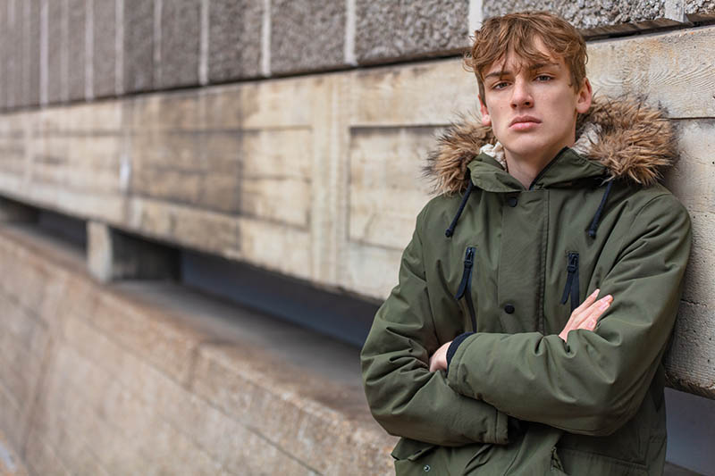 Young Man Male Teenager Boy Wearing Green Parka Young adult male teenager boy outside in an urban city wearing a green parka coat leaning against a wall